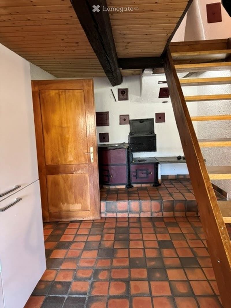 A narrow hallway with wooden stairs leading upwards, a wooden door on the left, and a small kitchen area visible at the end with a stove and sink.