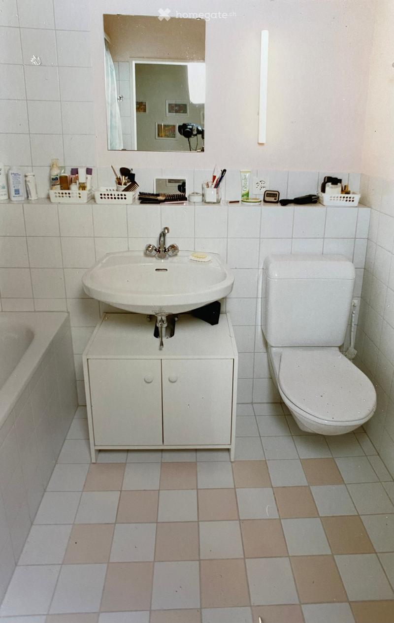 A small bathroom with a white sink, toilet, and mirror. The sink is mounted on a white cabinet with two doors. The floor is tiled with a mix of light and dark tiles. Various toiletries and personal care items are placed on the sink counter, including a to