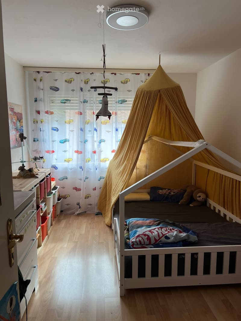 A cozy children's bedroom with a bunk bed, a tent-like play area, and colorful curtains featuring car designs. The room includes a storage unit with bins and a lamp on the nightstand.