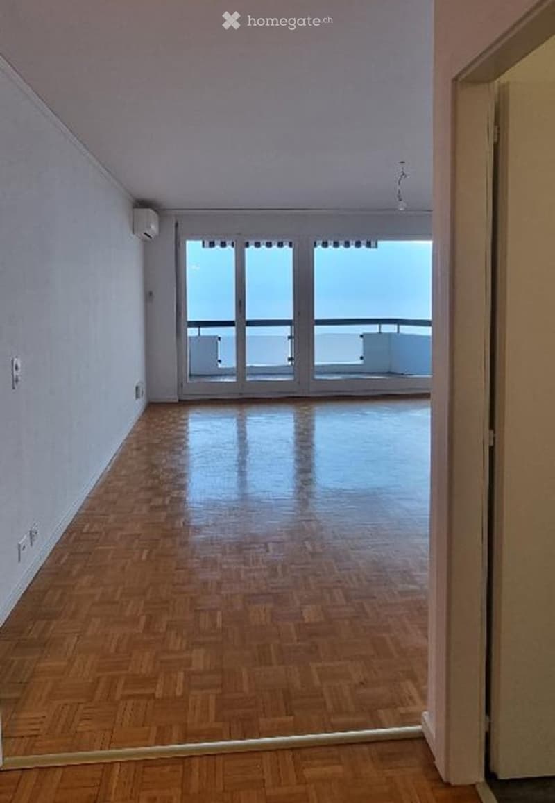 Empty room with wooden flooring, large windows offering a sea view, and access to a balcony.