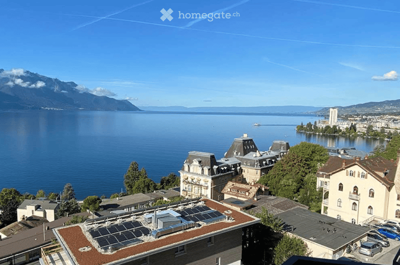 Aerial view of a lakeside town with historic buildings, solar panels on rooftops, and a scenic lake and mountain backdrop.