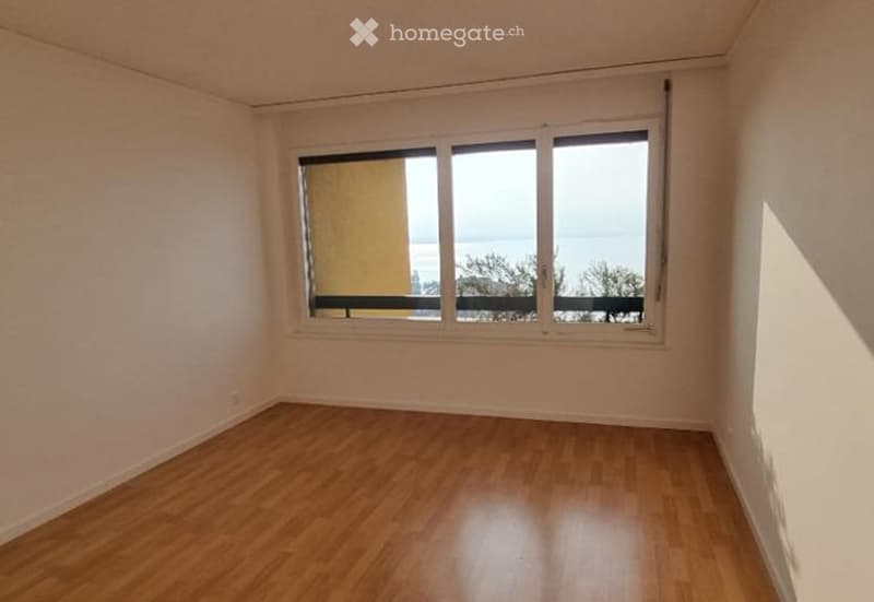 Empty room with wooden flooring and large window offering a view of the sea and surrounding area.
