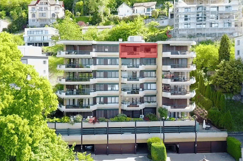 A multi-story residential building with balconies, large windows, and a prominent red accent wall. The structure is surrounded by greenery and includes a garage at the base.