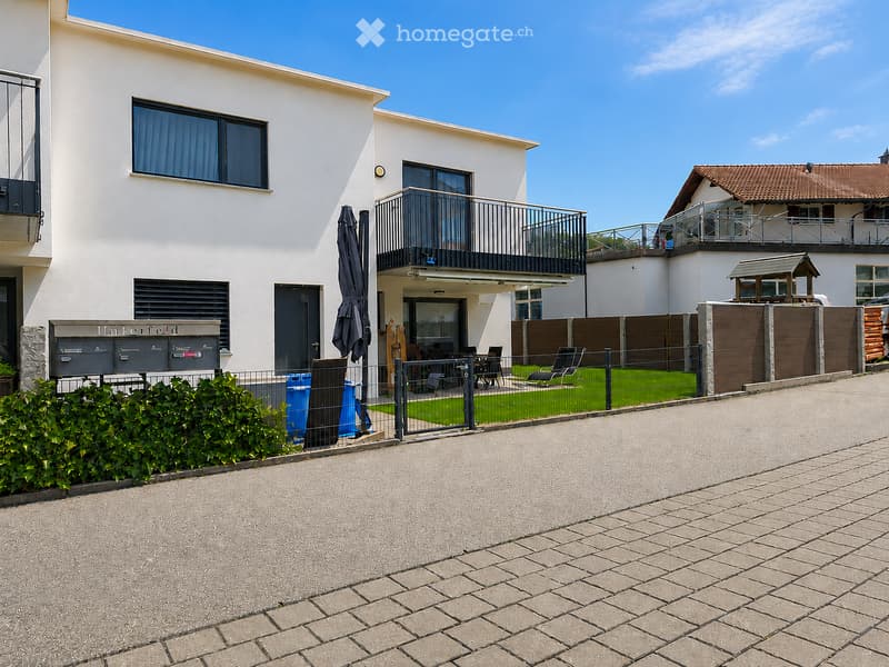 Modern two-story house with white exterior, large windows, balconies, and a fenced garden area with outdoor furniture.