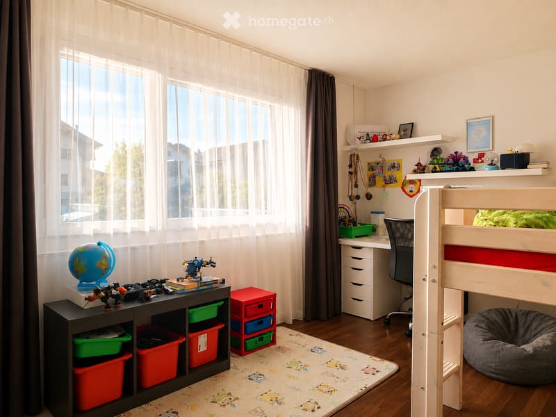 Brightly lit bedroom with a bunk bed, desk, and storage unit. Features a globe, toys, and colorful storage boxes. Large windows with sheer curtains and a view of greenery outside.