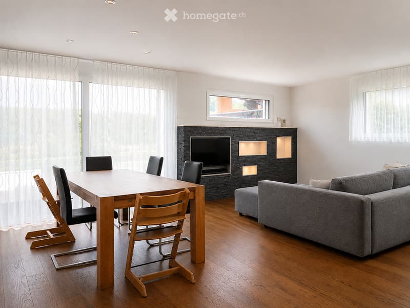 Modern living room with hardwood flooring, large windows, grey sectional sofa, wooden dining table with chairs, and a built-in entertainment unit with a TV and fireplace.