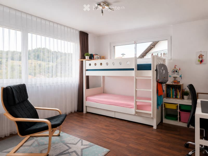 Bright bedroom with a white bunk bed, pink bedding, a black leather chair, a desk with a laptop, and a bookshelf with books and toys.
