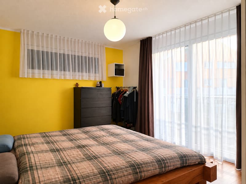 Modern bedroom with a large bed, plaid duvet, wooden nightstand, wardrobe, and large windows with sheer curtains.