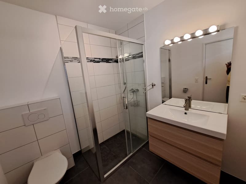 Modern bathroom with white tiles, a glass-enclosed shower, a double sink with a wooden vanity, and a large mirror with built-in lighting.
