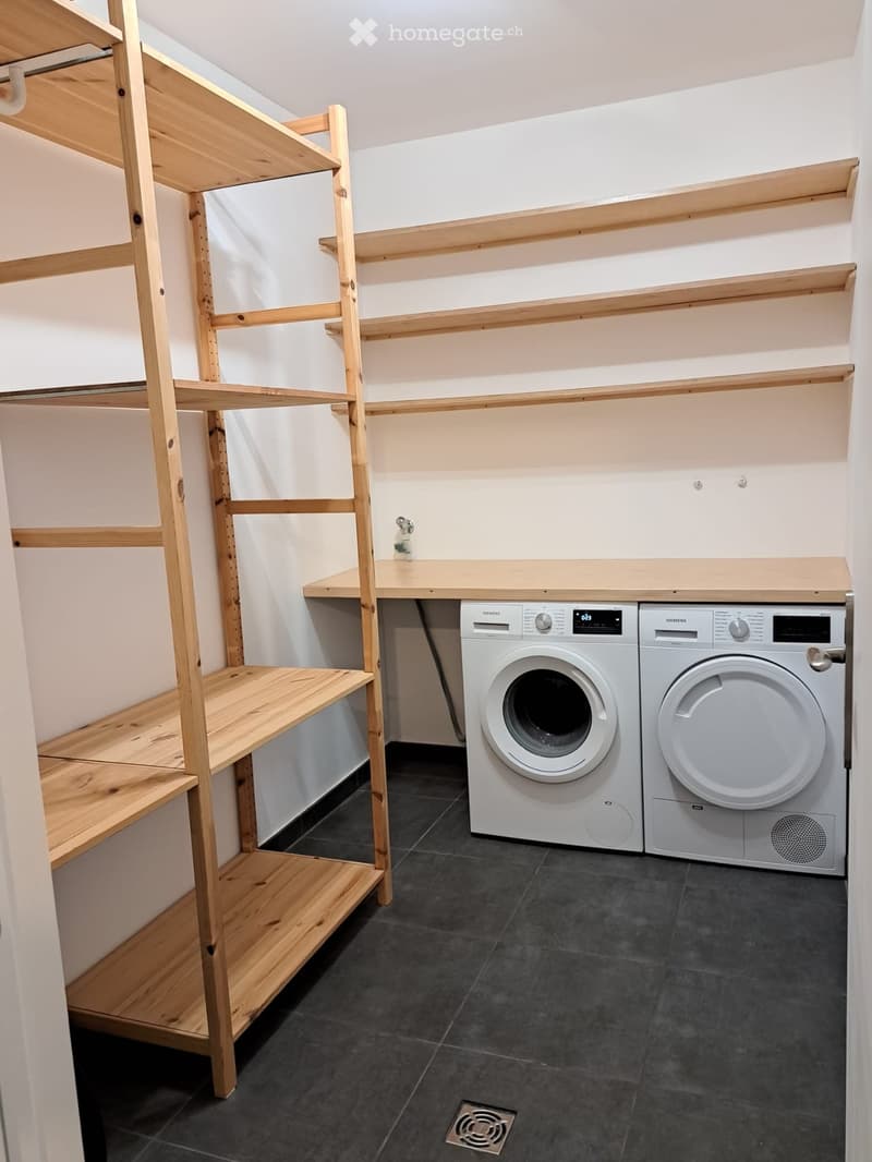Modern bathroom with dark tiled floor, white wooden shelves, and a washing machine and dryer unit.