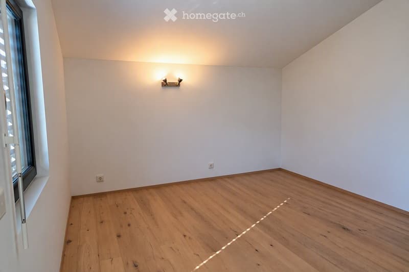 Empty room with wooden flooring, white walls, and a large window with blinds. The room features a modern wall-mounted light fixture and electrical outlets.