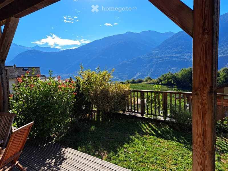 Balcony with wooden decking, wooden chairs, lush greenery, and a scenic mountain view.