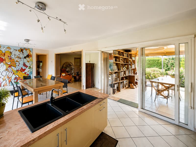 Modern kitchen with double sink, wooden countertop, dining table with chairs, bookshelf, and view to outdoor patio.