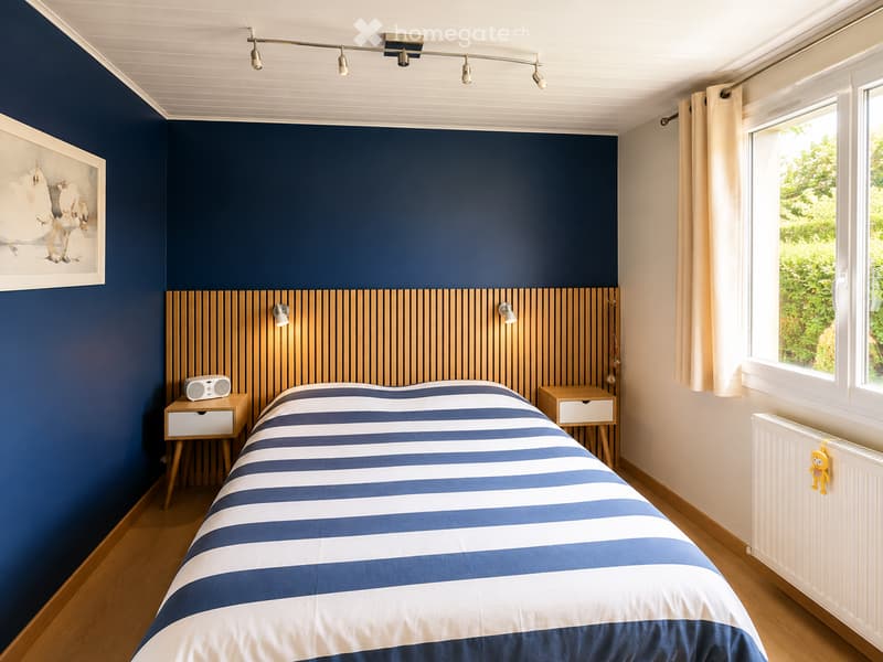 Modern bedroom with a blue accent wall, wooden headboard, and a bed with blue and white striped bedding. Features include a nightstand, a picture frame, and a window with curtains.