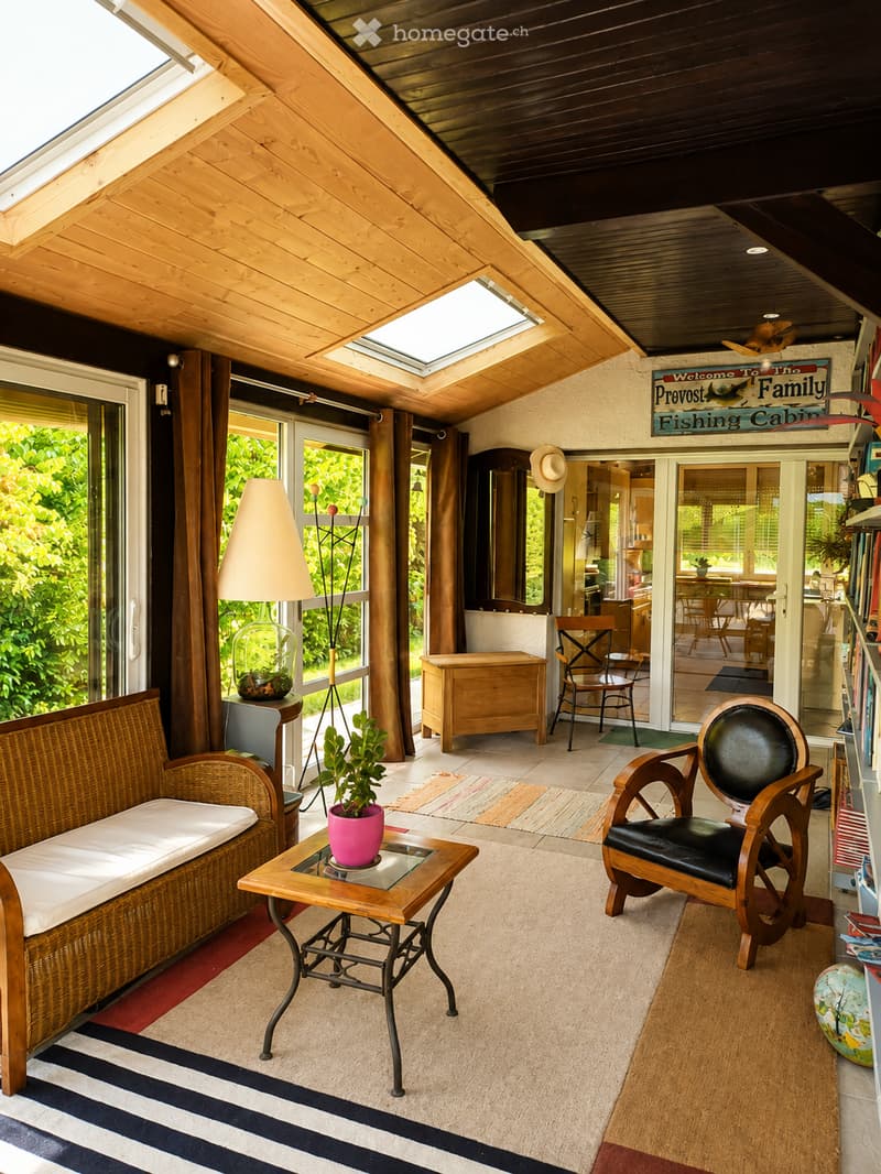 Bright living room with wooden ceiling, large windows, and skylight. Features a wicker sofa, glass coffee table with a pink plant, wooden armchair, floor lamp, and bookshelf.
