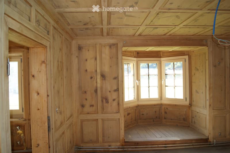 Empty wooden room with exposed beams, wooden walls, and multiple windows, showing natural light and wooden flooring.