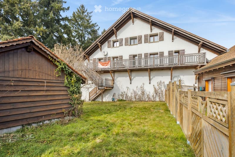 Two-story house with white exterior, wooden balcony, and wooden garage. The house features multiple windows with shutters and a well-maintained lawn in front.