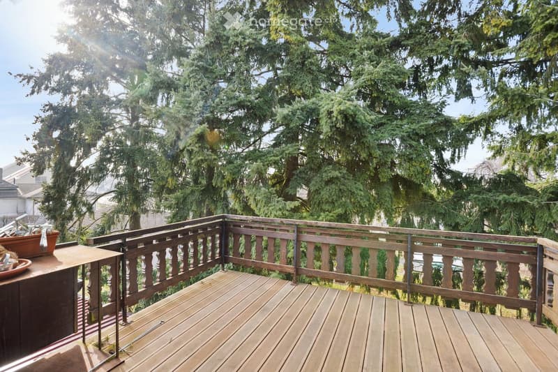Wooden balcony with railing, overlooking a lush green tree-filled area. The deck is made of light-colored wood planks, and the railing features vertical wooden balusters.
