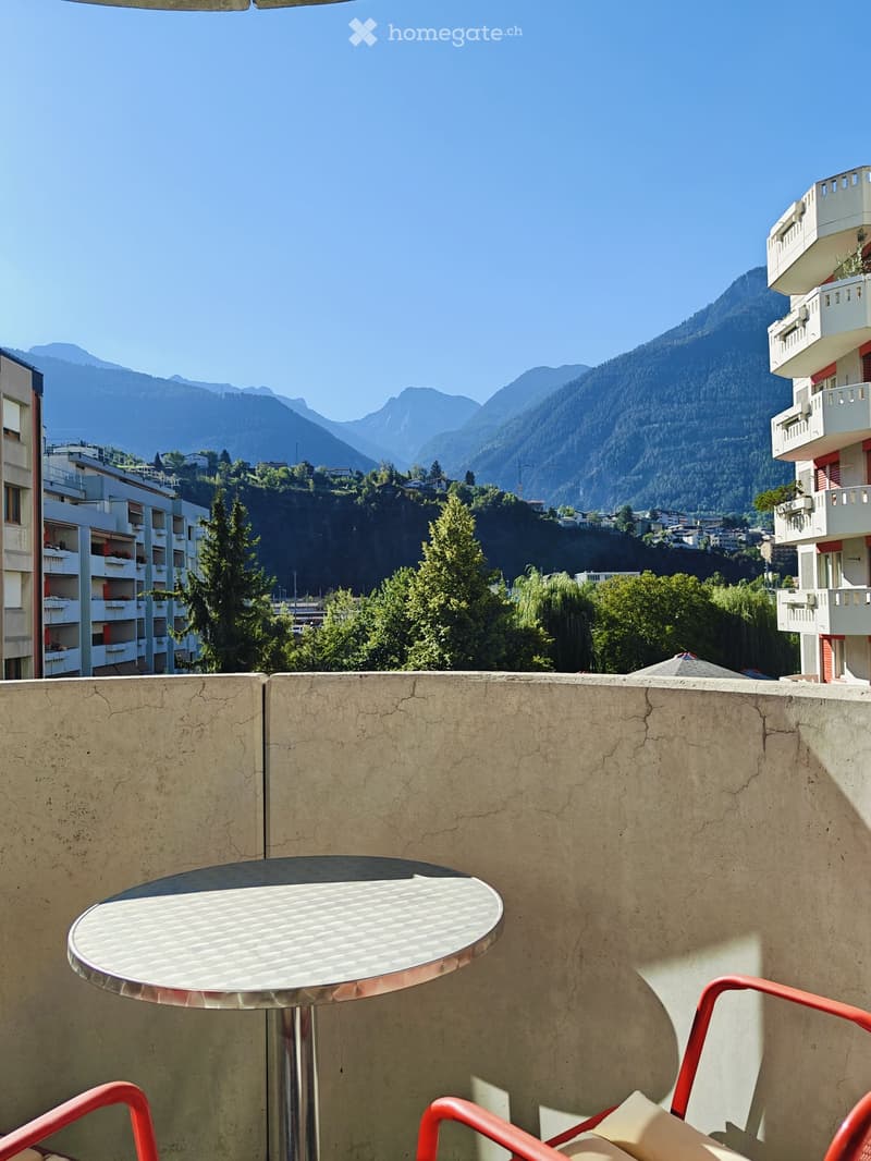 Balcony with a round metal table and red chairs, overlooking a scenic mountain view with surrounding buildings and greenery.