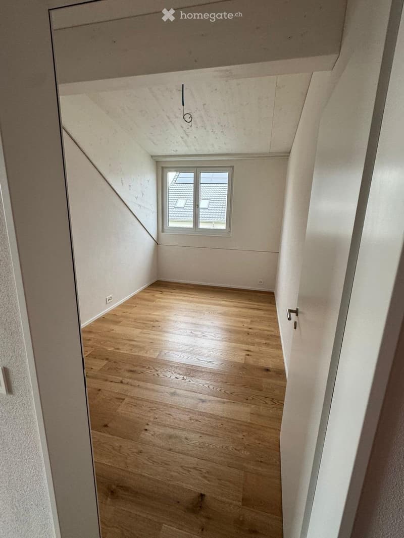 Empty attic space with wooden flooring, white walls, and a small window showing a roof view.