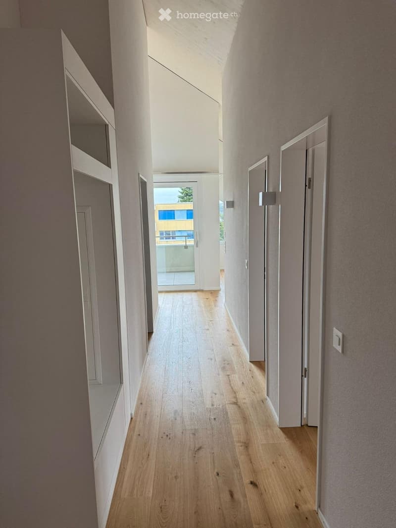 Modern hallway with light wooden flooring, white walls, and several doors leading to different rooms. Natural light streams in from the end of the corridor.