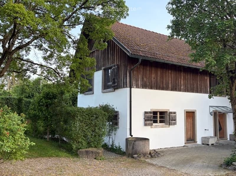 Traditional two-story house with white walls, wooden shutters, and a red-tiled roof, surrounded by greenery and a gravel driveway.