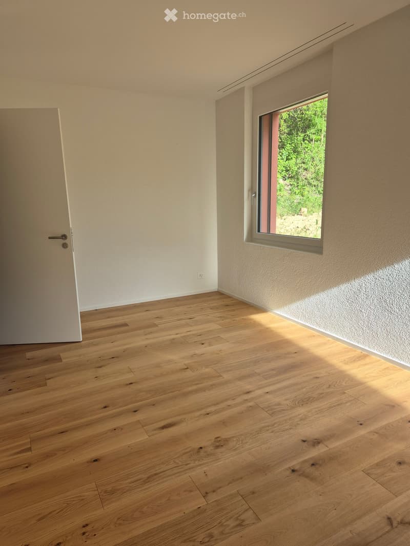 Empty room with light wooden flooring, white walls, and a large window showing greenery outside. The room has a door on the left side.