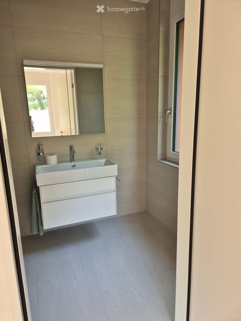 Modern bathroom with a white sink, mirror, and cabinet. The room features light-colored tiles and a window letting in natural light.