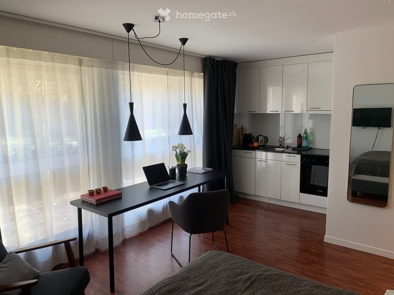 A modern, well-lit room with a sofa, a desk with a laptop, a kitchen area with cabinets, sink, and oven, and a mirror reflecting a television. The room features pendant lights, a large window with curtains, and a wooden floor.