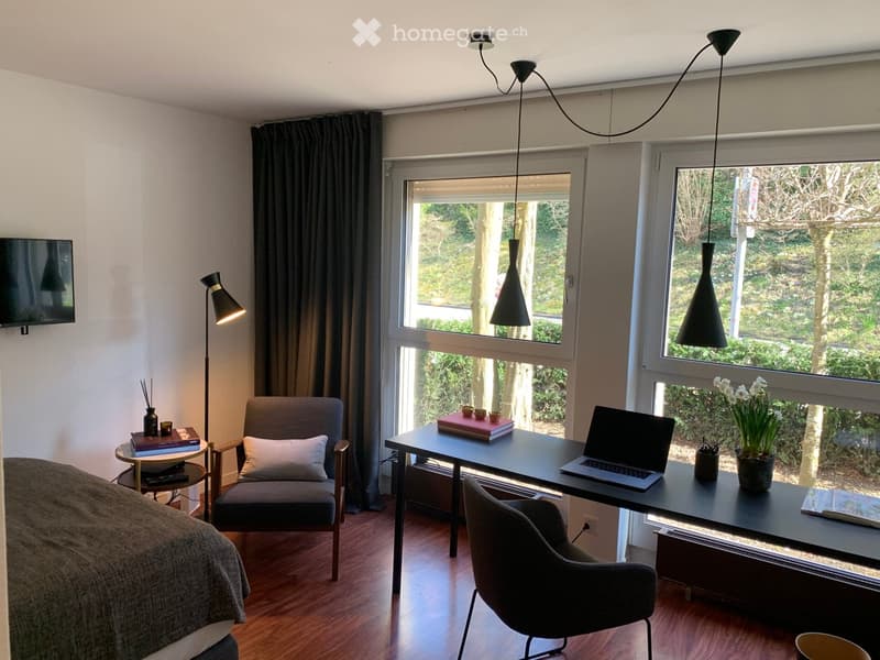 Cozy, well-lit room with a bed, desk, chair, laptop, and decorative items. Large windows offer a view of greenery outside.