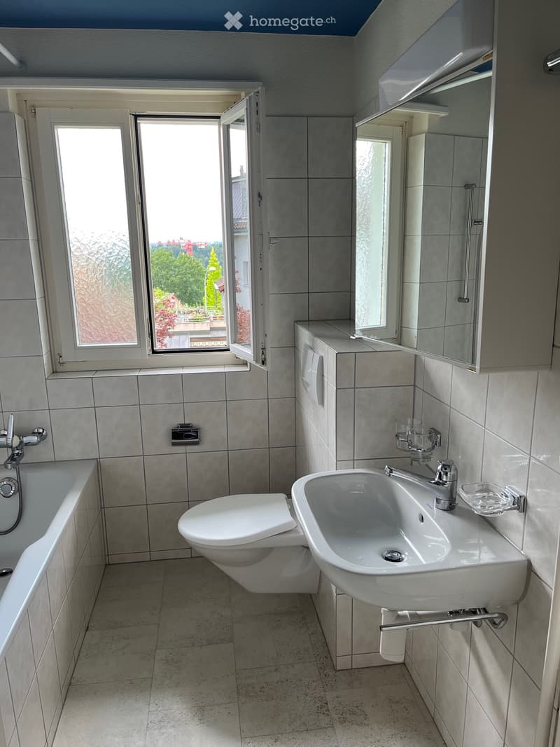 Modern bathroom with a bathtub, toilet, sink, and mirrored cabinet. Large window offers a view of greenery outside.
