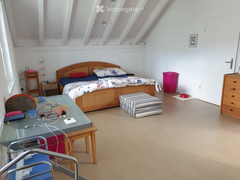 A cozy bedroom with a wooden bed, nightstand, glass table, and a small chair. The room features a slanted ceiling and a small stool near the bed.