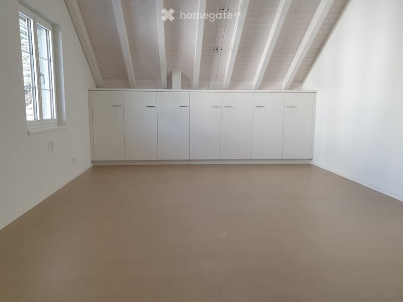 Empty room with white walls, exposed wooden beams on the ceiling, a large window, and built-in white cabinets.