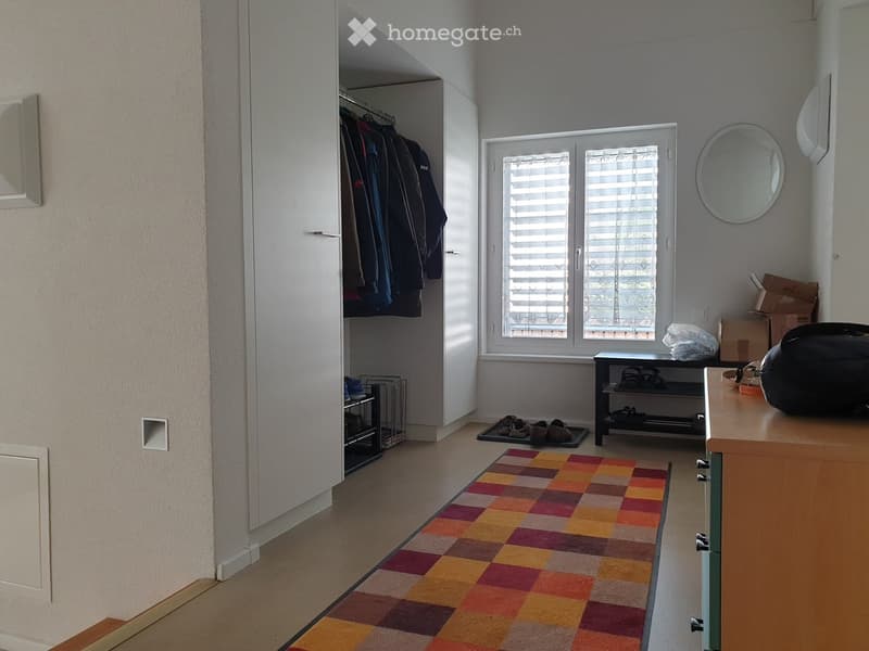 The image shows a well-lit hallway or entryway with a colorful checkered rug, a white wardrobe with hanging clothes, a round mirror on the wall, a shoe rack, and a small table with items on it.