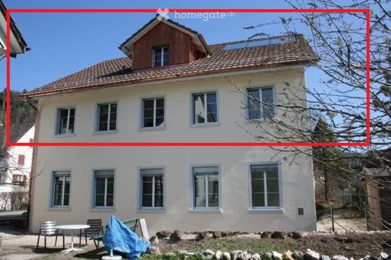 Two-story house with a beige facade, multiple windows, and a tiled roof. The house has a small garden area in front with a table and chairs.