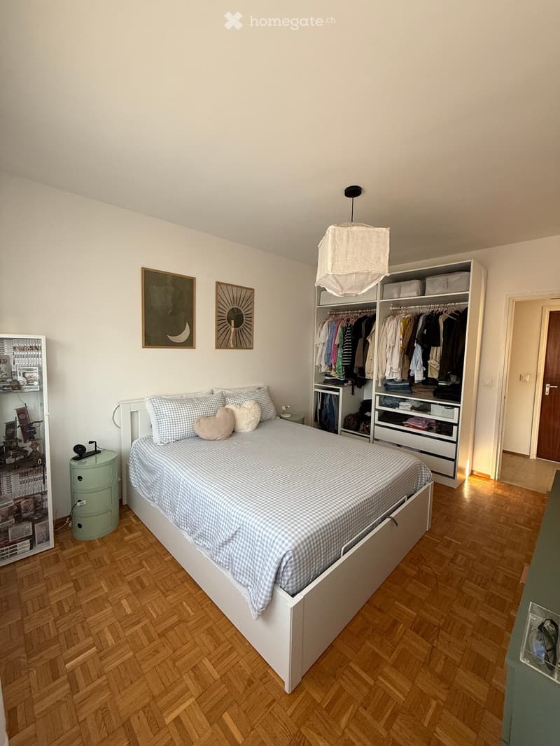 Cozy bedroom with a white bed, checkered bedding, pillows, a nightstand, a wardrobe with clothes, and a bookshelf filled with books.