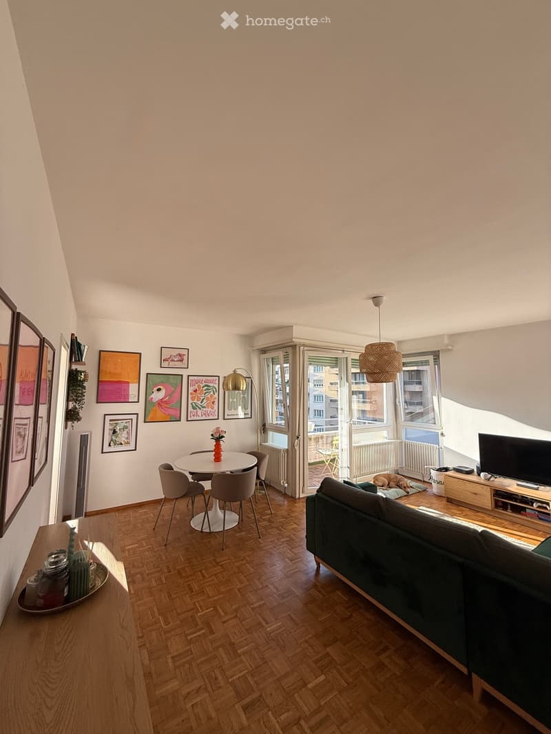 Living room with wooden flooring, a grey sofa, a white dining table with four chairs, a television, a floor lamp, and several picture frames on the wall. A dog is lying on a mat near the balcony door.
