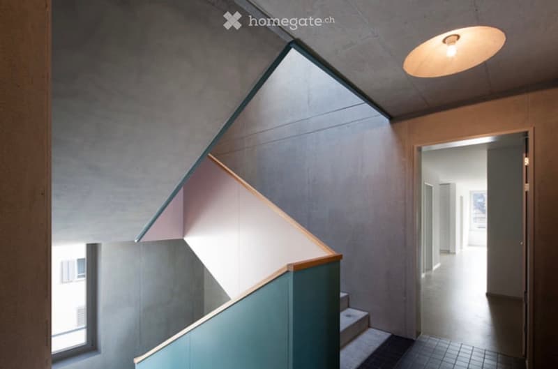 Modern hallway with a staircase featuring a geometric railing, a pendant light fixture, and a view into an adjacent open space.
