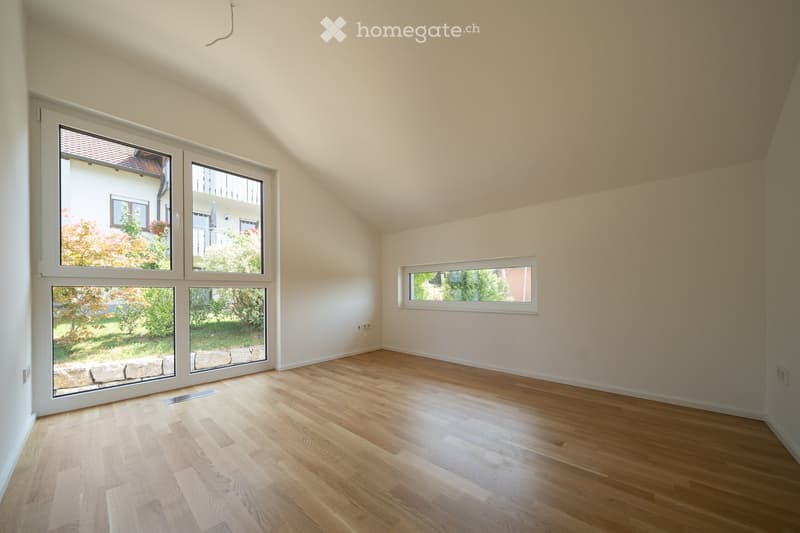 Empty room with wooden flooring, white walls, and large windows offering views of the garden and neighboring house.