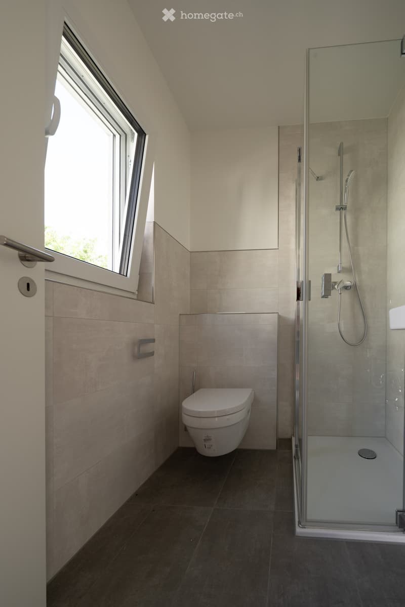 Modern bathroom with a toilet, glass-enclosed shower, and a window providing natural light.
