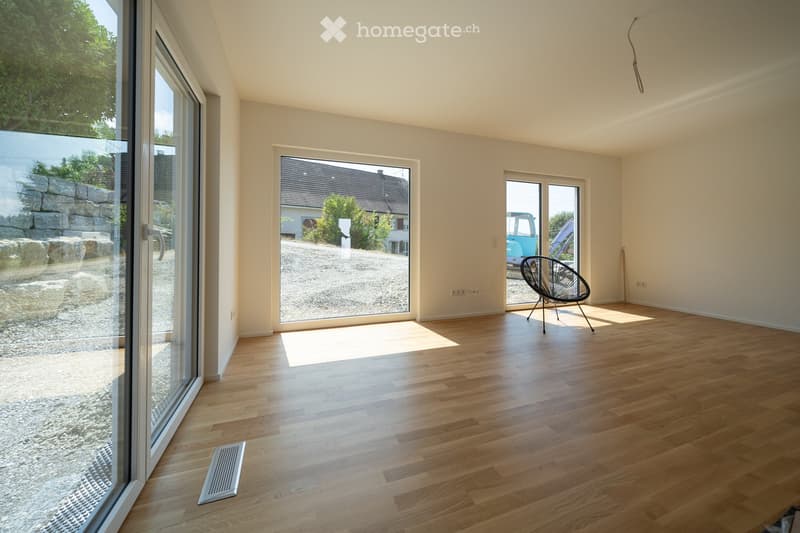 Empty room with wooden flooring, large windows, and a single modern chair near the door.