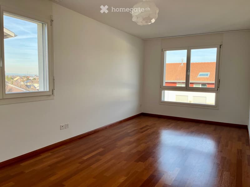 Empty room with wooden flooring, white walls, and large windows offering a view of the city and rooftops.