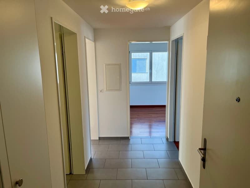 A clean, well-lit hallway with tiled flooring, white walls, and several doors leading to different rooms. The hallway features a ceiling light and a visible electrical outlet on one wall.