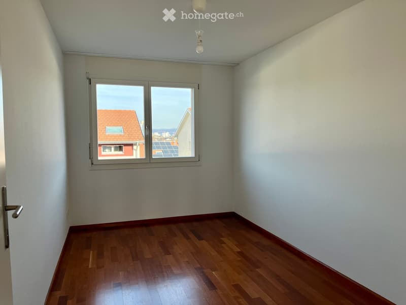 Empty room with wooden flooring, white walls, and a large window offering a view of neighboring buildings and solar panels.
