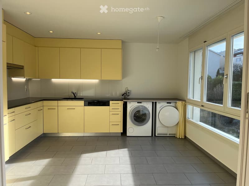 Bright kitchen with light yellow cabinets, stainless steel sink, dishwasher, washing machine, and dryer. Large windows provide natural light.