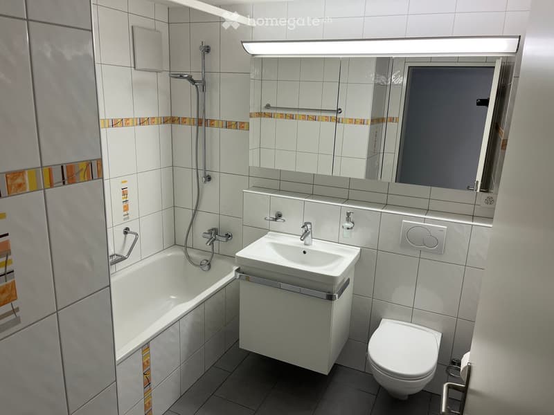 Modern bathroom with white tiles, bathtub, sink, toilet, and large mirror above the sink.