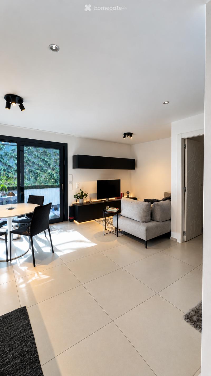 Modern living room with a grey sofa, white dining table, chairs, TV, and a small plant on a side table. Large sliding glass door leads to a balcony.