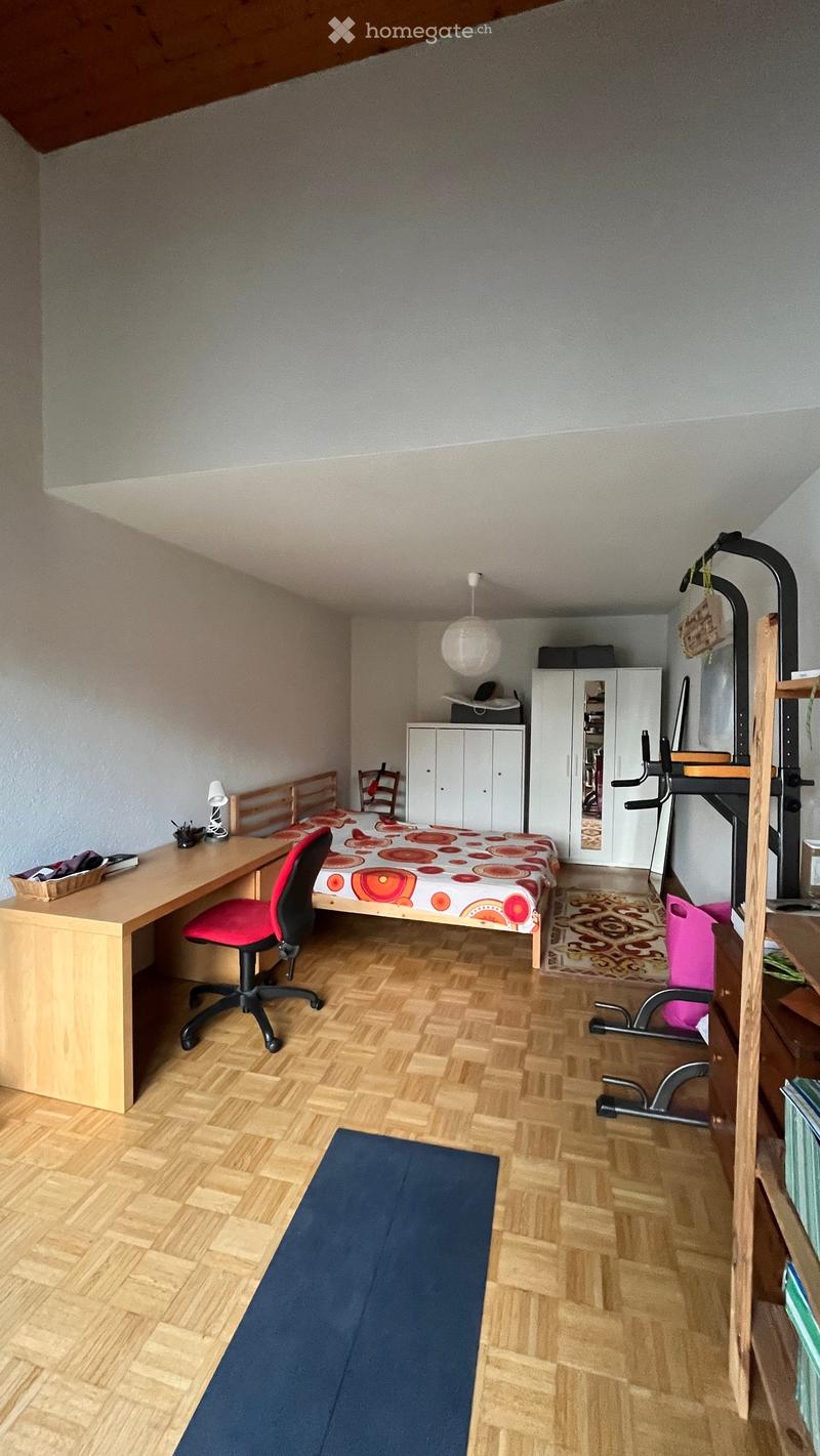 A small, furnished bedroom with a wooden desk, red chair, bed with a colorful bedspread, white wardrobe, and exercise bike in the corner.