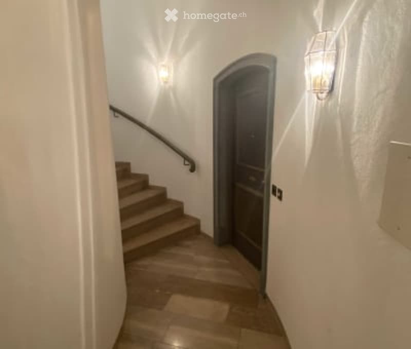 A well-lit hallway with a wooden staircase leading upwards, featuring a dark wooden door on one side and wall-mounted lighting fixtures.