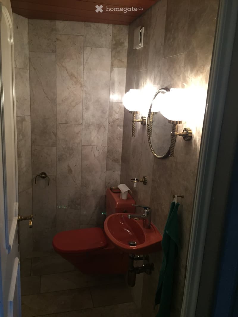 A small bathroom with a red toilet and sink, marble tiles, a wall-mounted light fixture, and a green towel hanging on a rack.